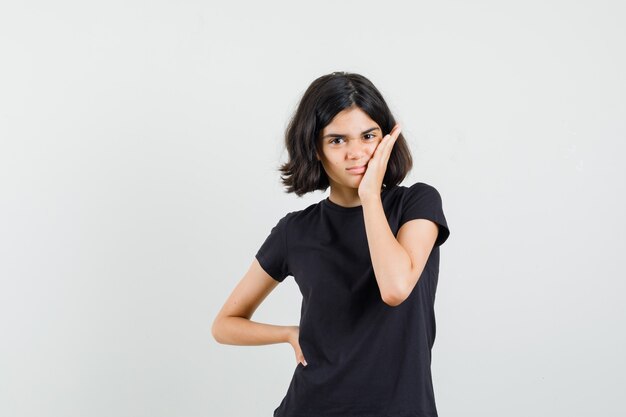 Little girl holding palm on cheek in black t-shirt and looking cute. front view.