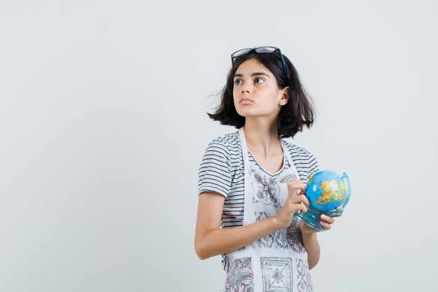 Little girl holding school globe in t-shirt, apron and looking pensive.