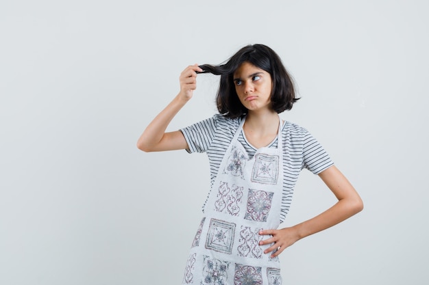 Little girl holding strand of her hair in t-shirt, apron and looking pensive ,