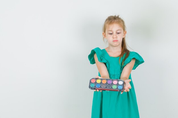 Little girl holding watercolor paints with brush in green dress and looking serious