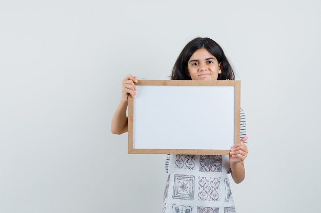 Little girl holding white frame in t-shirt, apron and looking confident ,