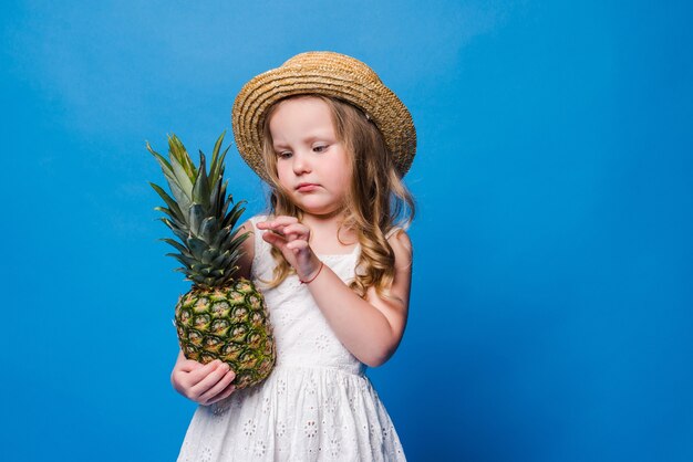 Little girl holds whole pineapple on blue wall with copy space