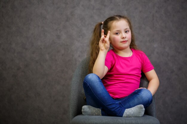 Маленькая девочка в розовой футболке сидит на сером стуле. эмоциональное фото ребенка