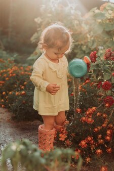Маленькая девочка в желтом платье и резиновых сапогах поливает цветы в саду маленький помощник