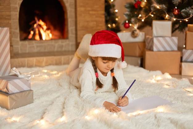 Маленькая девочка в красной рождественской шляпе пишет письмо деду морозу, очаровательному ребенку, лежащему на полу у камина, в белом теплом джемпере, лежащему на полу на мягком ковре.