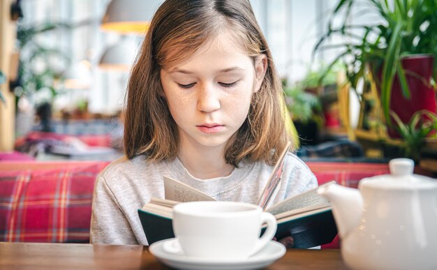 A little girl is reading a book while sitting in a cafe with a cup of tea