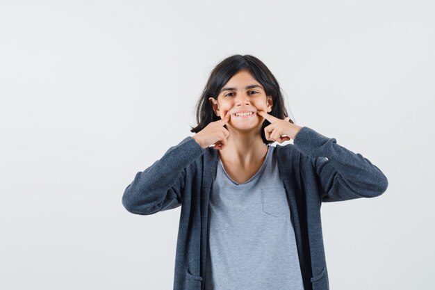 Little girl keeping fingers on cheeks in t-shirt, jacket and looking cheery.