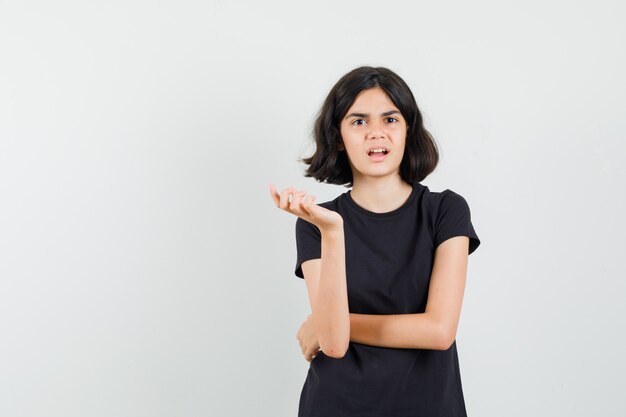 Little girl keeping hand in questioning manner in black t-shirt and looking confused , front view.