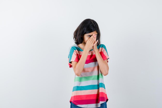 Маленькая девочка держит руки на лице в футболке и выглядит обеспокоенной, вид спереди.