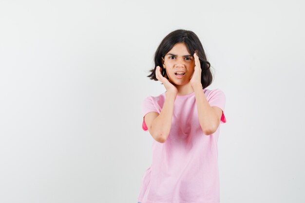 ピンクのTシャツで顔の近くに手を保ち、不満を探している少女。正面図。