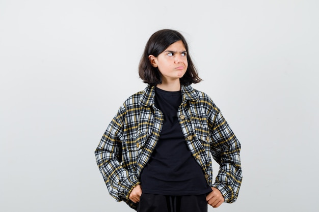 Little girl keeping hands in pockets in t-shirt, jacket and looking thoughtful. front view.