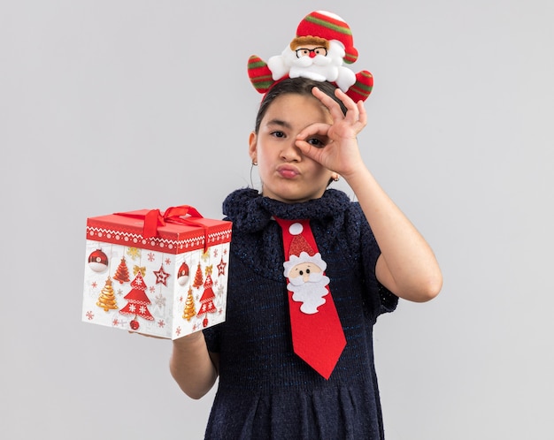 ニットのドレスを着た少女は、頭に面白いクリスマスの縁が付いた赤いネクタイを着て、クリスマスプレゼントを持って幸せそうに見えてポジティブに見えます。