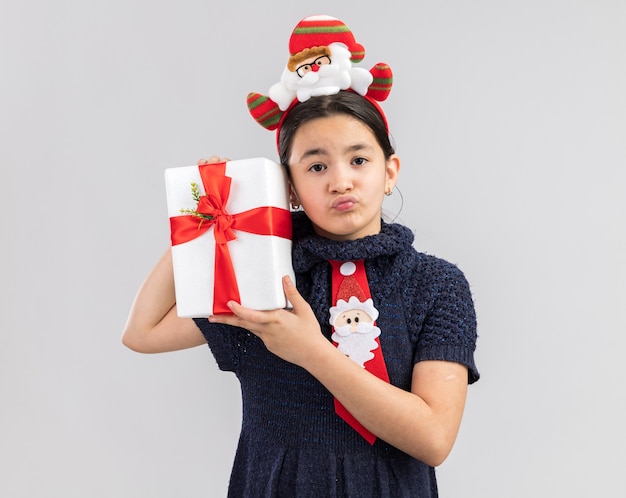 Little girl in knit dress wearing red tie with funny christmas rim on head holding christmas present looking confused with sad expression 