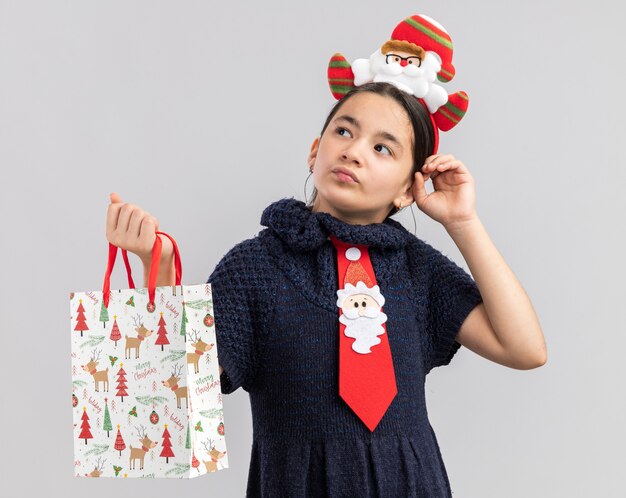 Little girl in knit dress wearing red tie with funny christmas rim on head holding paper bag with christmas gift looking aside puzzled 
