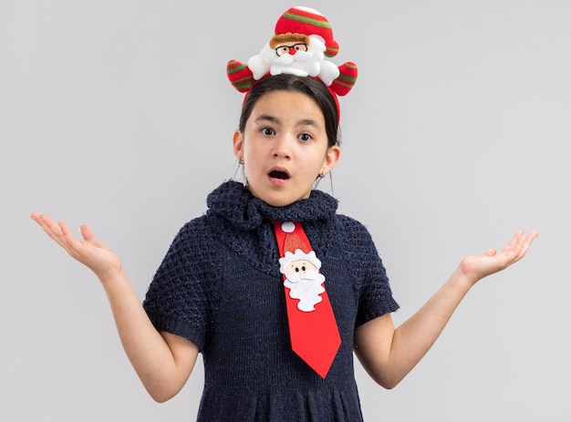 Little girl in knit dress wearing red tie with funny christmas rim on head looking amazed with arms raised 