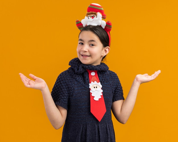 Маленькая девочка в вязаном платье в красном галстуке с забавным рождественским ободком на голове выглядит счастливой и веселой, улыбаясь, разводя руки в стороны