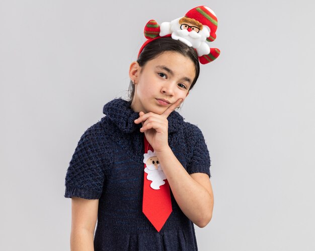 의아해 보이는 머리에 재미있는 크리스마스 테두리와 빨간 넥타이를 착용하는 니트 드레스에 어린 소녀