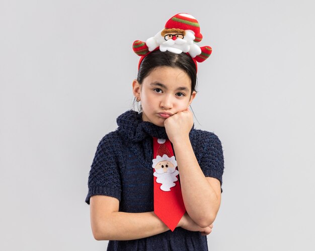 Little girl in knit dress wearing red tie with funny christmas rim on head looking tired and bored 