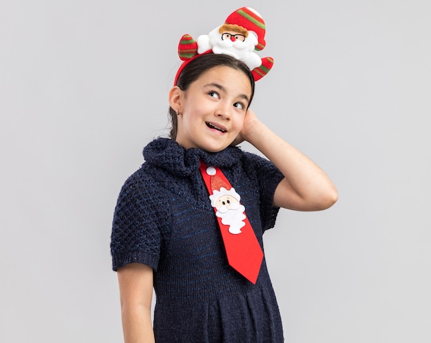 행복하고 긍정적 인 찾고 머리에 재미있는 크리스마스 테두리와 빨간 넥타이를 입고 니트 드레스에 어린 소녀
