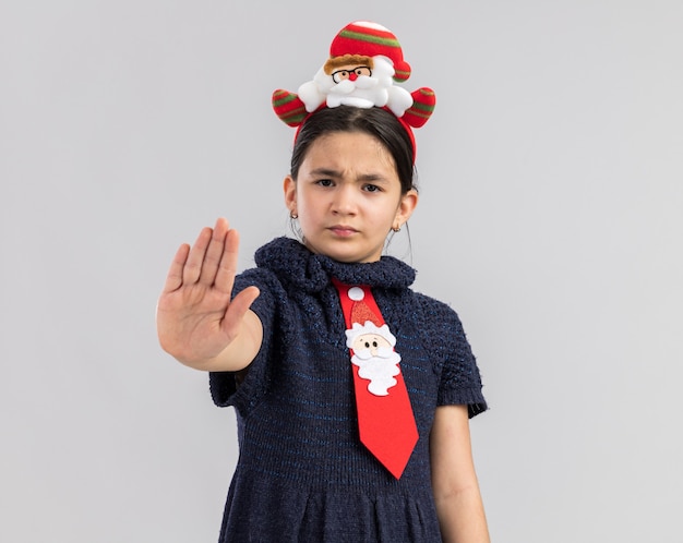 Little girl in knit dress wearing red tie with funny christmas rim on head looking with serious face making stop gesture with had 