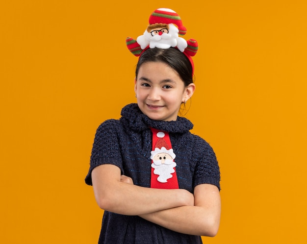 Маленькая девочка в вязаном платье в красном галстуке с забавным рождественским ободком на голове смотрит с улыбкой на лице, скрестив руки