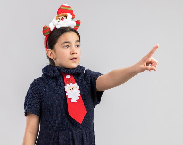 Little girl in knit dress wearing red tie with funny christmas rim on head pointing with index finger at something aside worried 