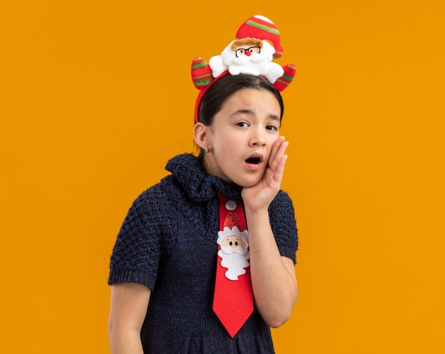 Little girl in knit dress wearing red tie with funny christmas rim on head whispering with hand near mouth 