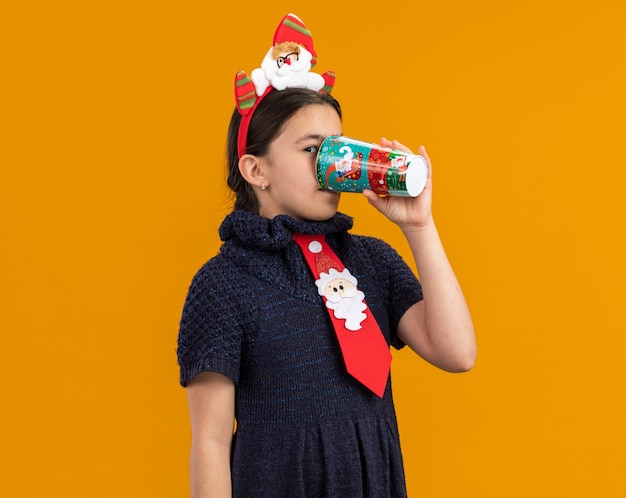 Little girl in knit dress wearing red tie with funny rim on head drinking from colorful paper cup happy and positive 