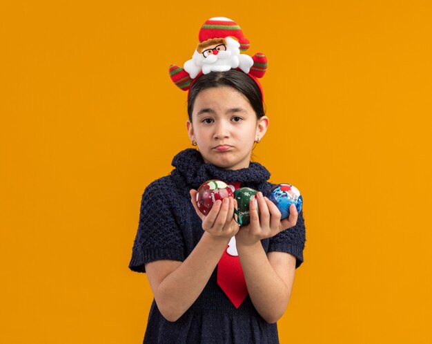 Little girl in knit dress wearing red tie with funny rim on head holding christmas balls looking with sad expression 