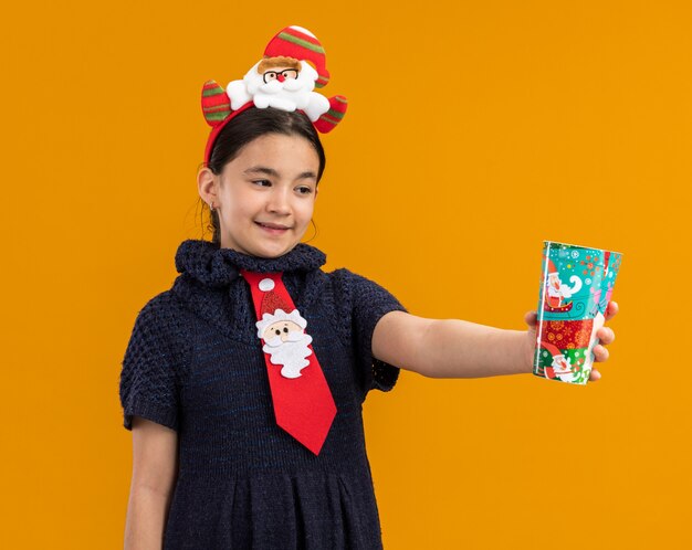 Little girl in knit dress wearing red tie with funny rim on head holding colorful paper cup happy and positive smiling 