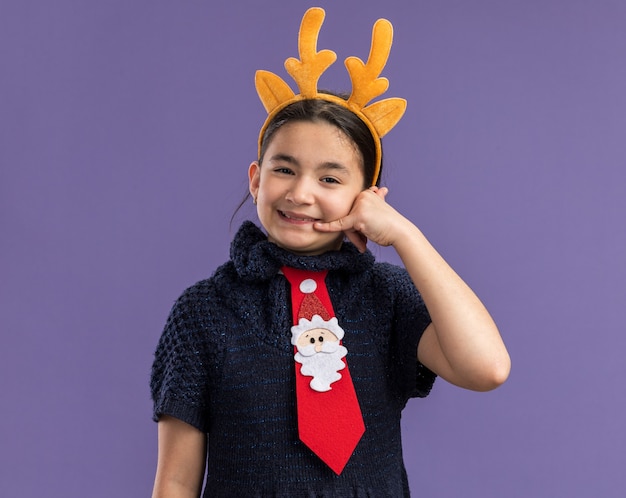 Little girl in knit dress wearing  red tie with funny rim with deer horns on head   happy and positive making call me gesture smiling  standing over purple wall