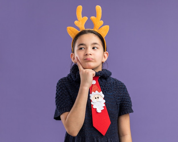 Little girl in knit dress wearing red tie with funny rim with deer horns on head looking aside puzzled 