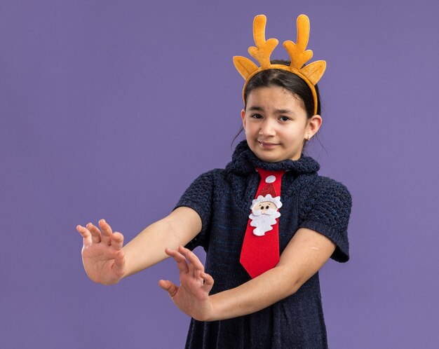 Little girl in knit dress wearing red tie with funny rim with deer horns on head  looking scared making defense gesture with hands  standing over purple wall