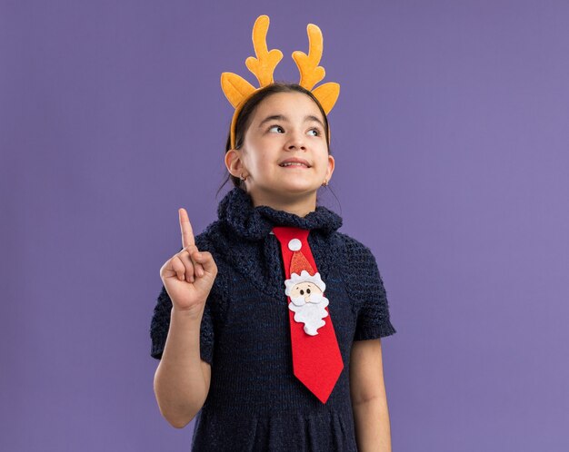 Little girl in knit dress wearing red tie with funny rim with deer horns on head  looking up with smile on surprised face showing index finger having new idea standing over purple wall