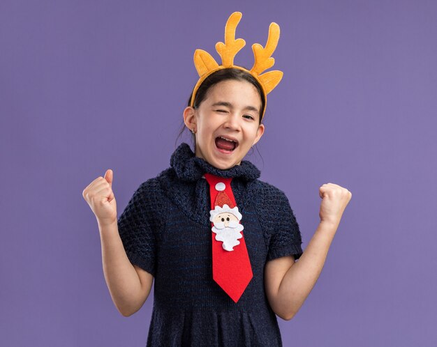 Little girl in knit dress wearing  red tie with funny rim with deer horns on head  screaming happy and excited clenching fists  standing over purple wall