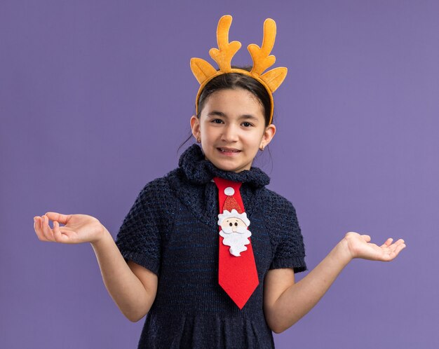 Little girl in knit dress wearing  red tie with funny rim with deer horns on head   smiling confused spreading arms to the sides  standing over purple wall