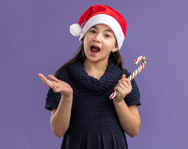 Маленькая девочка в вязаном платье в шляпе санта-клауса с конфетой в руках, счастливая и удивленная, стоя над фиолетовой стеной