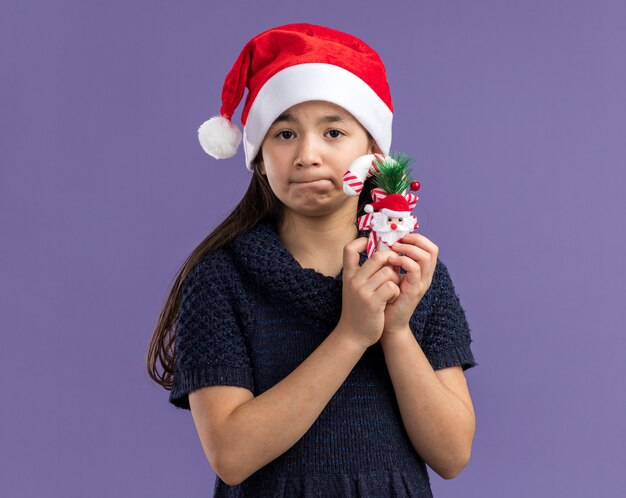 보라색 벽 위에 서있는 얼굴에 슬픈 표정과 혼동 크리스마스 사탕 지팡이를 들고 산타 모자를 쓰고 니트 드레스에 어린 소녀
