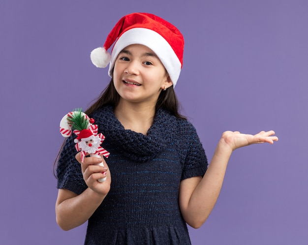 보라색 벽 위에 행복하고 밝은 서 얼굴에 미소로 크리스마스 사탕 지팡이를 들고 산타 모자를 쓰고 니트 드레스에 어린 소녀