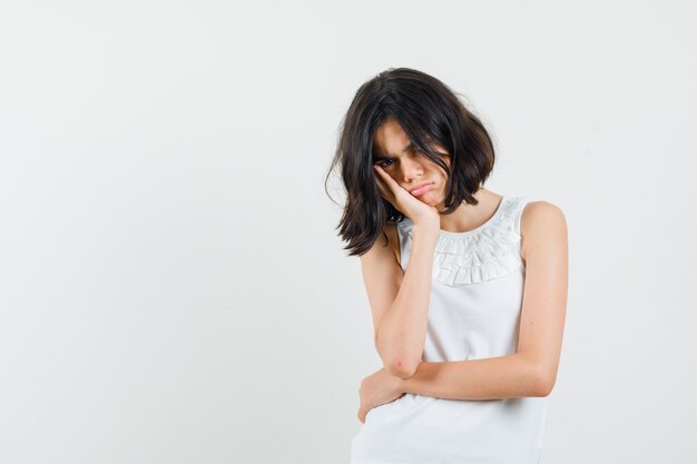 Little girl leaning cheek on raised palm in white blouse and looking offended , front view.