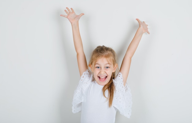 Little girl lifting both hands up in white t-shirt and looking cheery