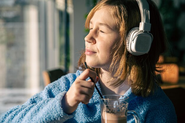 小さな女の子はヘッドフォンで音楽を聴き、晴れた朝にココアを飲みます