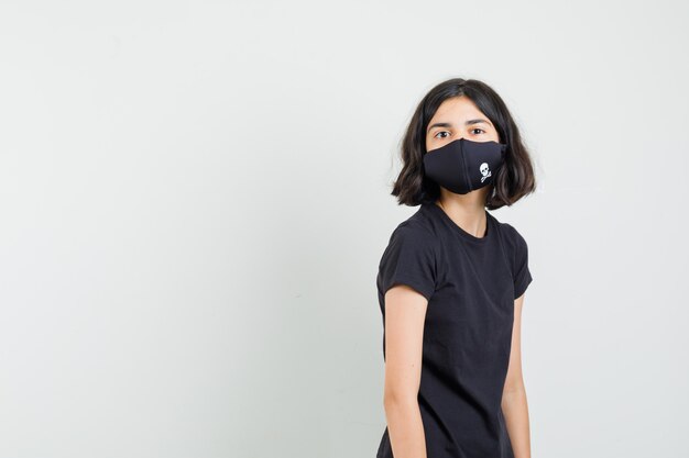Little girl looking at front in black t-shirt, mask and looking sensible , front view.