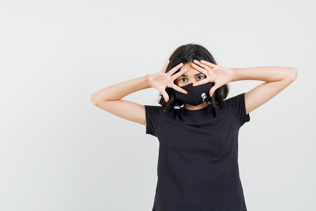 Little girl looking through fingers in black t-shirt, mask , front view.