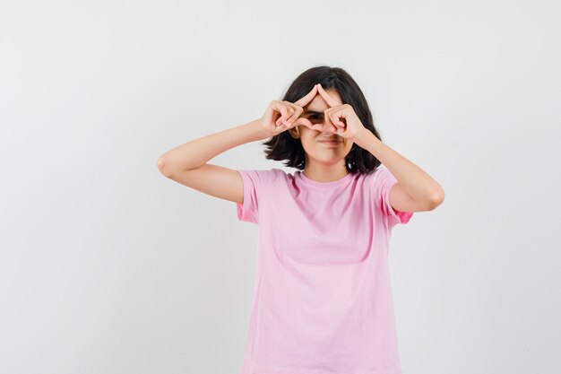 Маленькая девочка смотрит сквозь пальцы в розовой футболке и выглядит веселой. передний план.