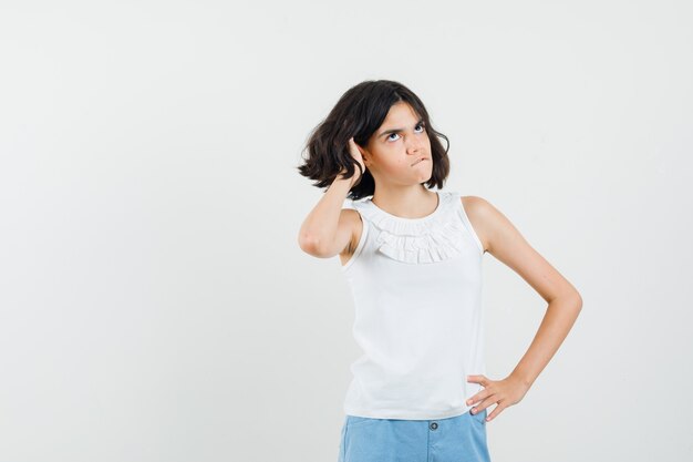 Little girl looking up, biting lip, holding hand in hair in white blouse, shorts front view.