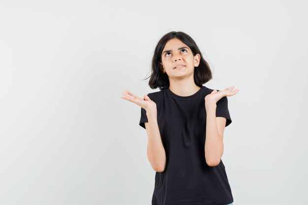 Little girl looking up, raising hands in black t-shirt and looking curious , front view.