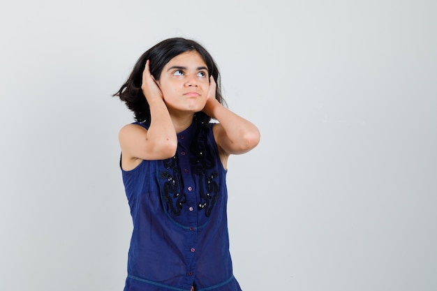 Little girl looking up while holding hands on ears in blue blouse.