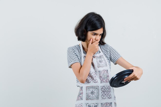 Little girl looking at wall clock in t-shirt, apron and looking forgetful.