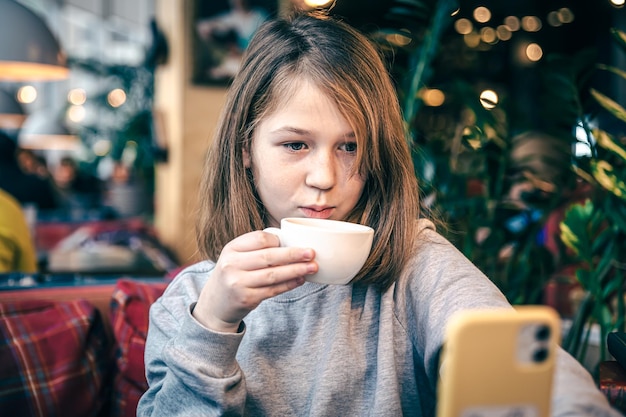 小さな女の子がカフェに座ってスマートフォンの画面を見ます